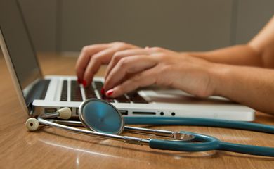 Médica digita no computador, tendo seu estetoscópio ao lado. Foto: National Cancer Institute/Unsplash